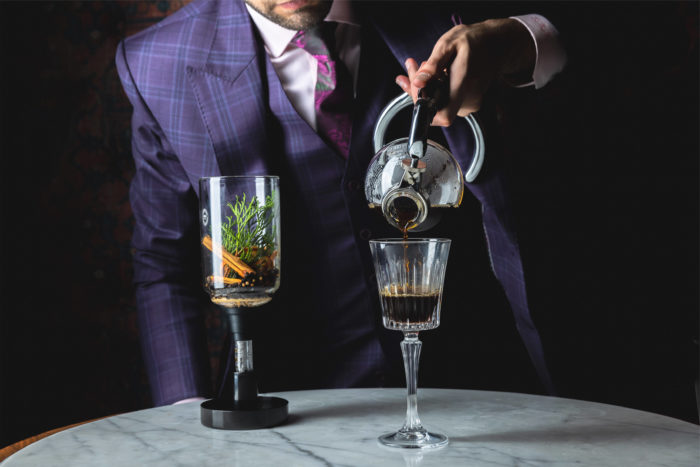 A person in a purple plaid suit pours a dark liquid from a siphon into a tall glass goblet.