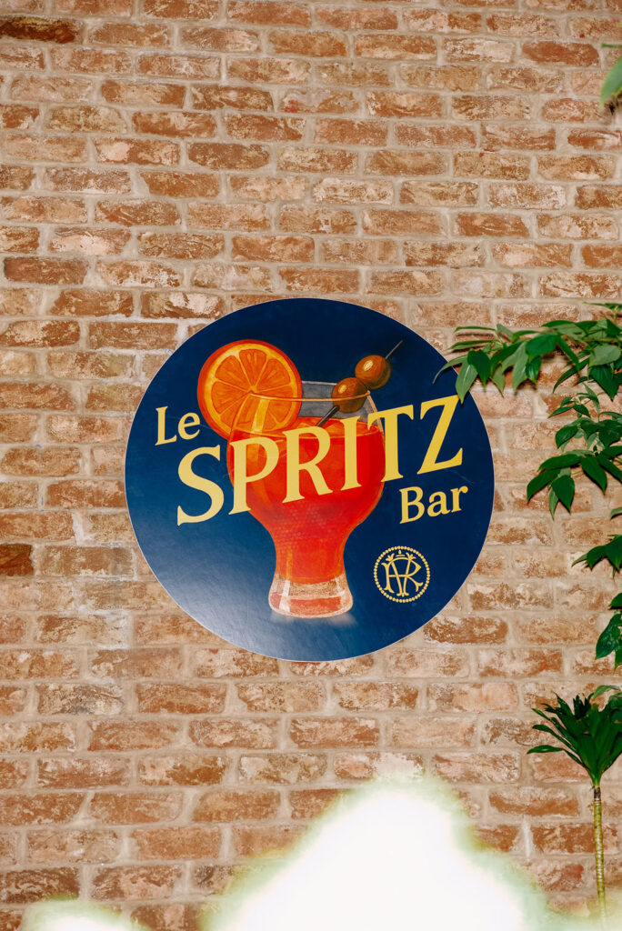 Round sign on a brick wall reading "Le Spritz Bar" with an illustration of a red spritz cocktail garnished with an orange slice and two olives.