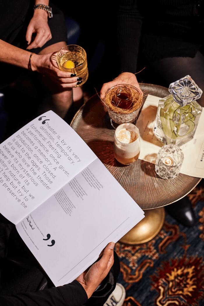 Overhead view of cocktails on a round table with hands holding drinks and an open book on a colorful carpet.