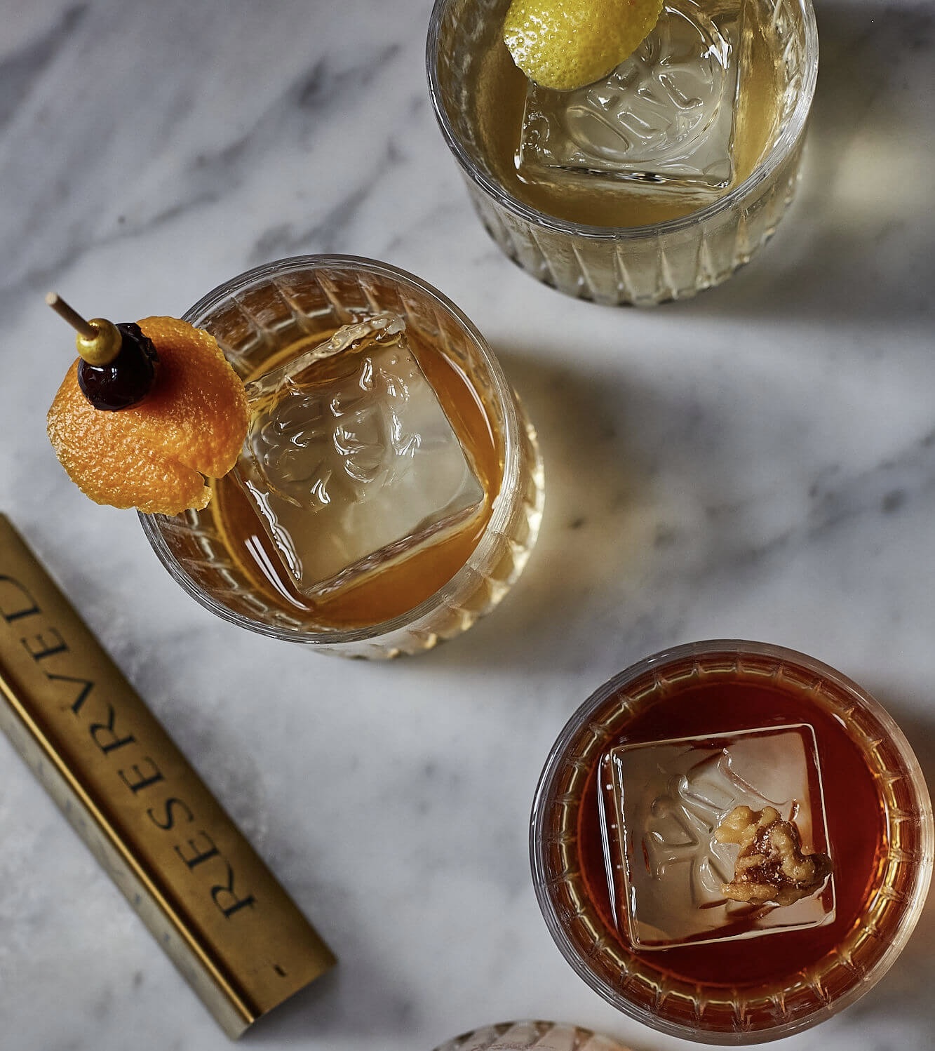 Overhead view of four cocktails with large ice cubes and garnishes on a marble surface.