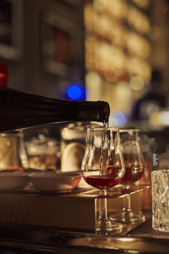 A bottle pouring red wine into a glass at a dimly lit bar with blurred bottles in the background.