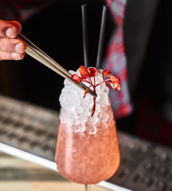 Close-up of a pink cocktail topped with crushed ice, garnished with red floral sprigs using tweezers, with a bartender in plaid in the background.
