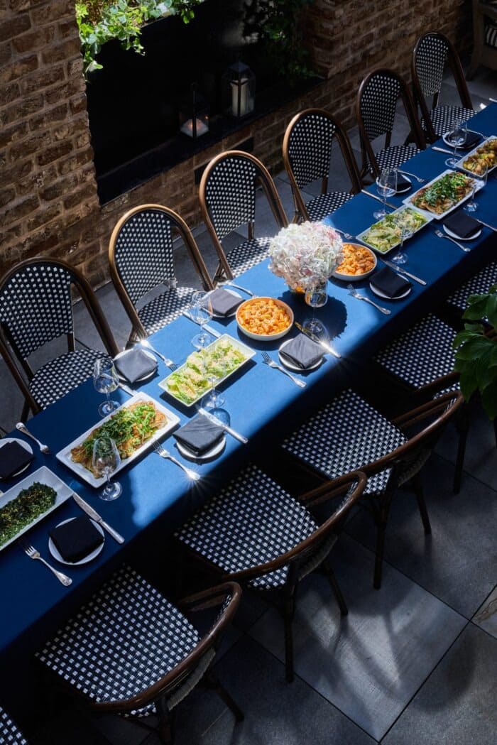 Elegant long dining table with blue tablecloth, set for a group meal in a stylish indoor-outdoor venue. The table features black-and-white woven chairs, glassware, utensils, and several dishes including pasta and salads, with a central bouquet of white and pink flowers. Brick wall and greenery add ambiance to the space.