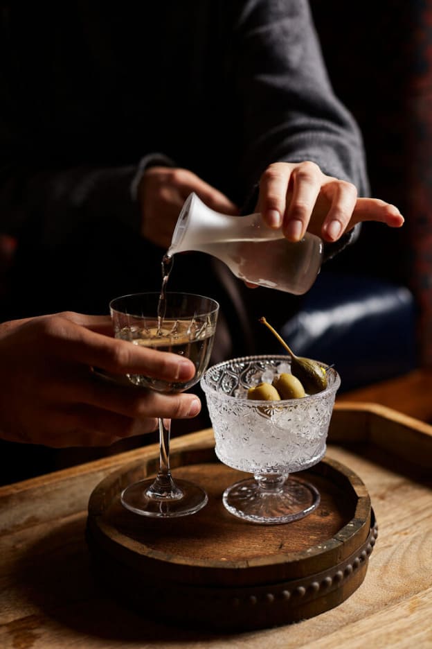 Hands pouring a clear spirit into a coupe glass beside a chilled martini glass with olives.