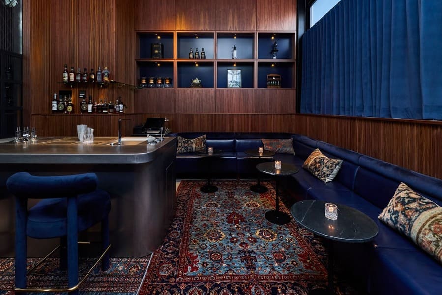 Elegant cocktail lounge with blue velvet seating, dark wood walls, and backlit liquor shelves.