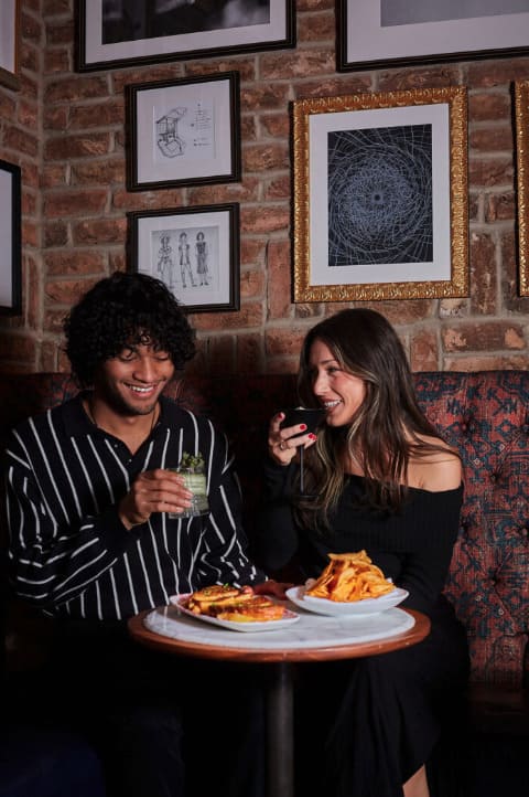 A smiling couple sits at a cozy restaurant table with cocktails and shared plates of food, including fries and a sandwich, surrounded by brick walls and framed artwork.