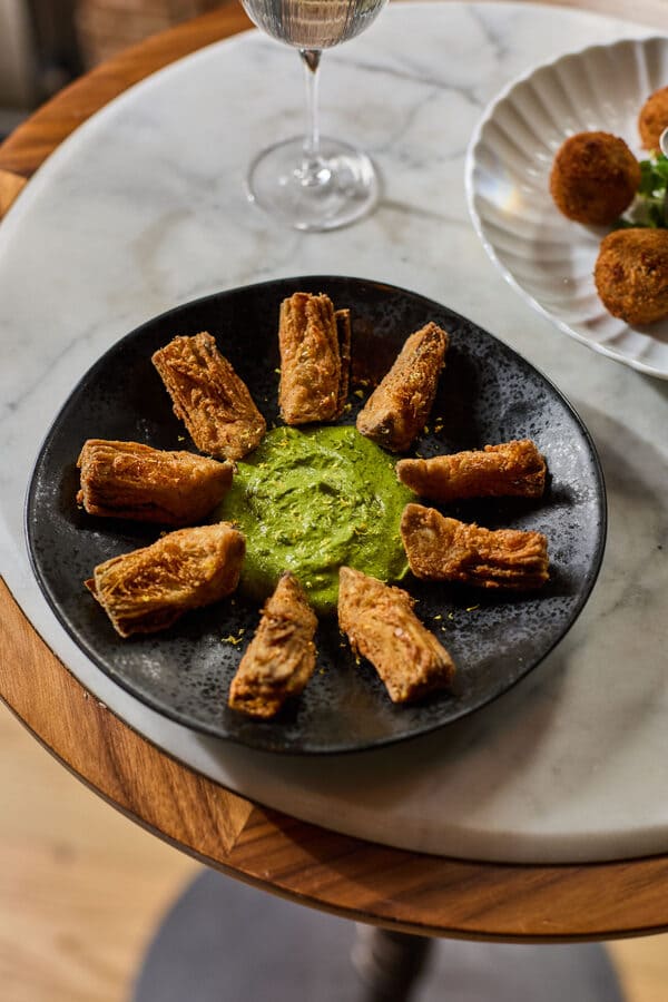 Golden fried vegetable rolls arranged in a circle around a bowl of bright green dipping sauce on a black plate placed on a marble-topped table.