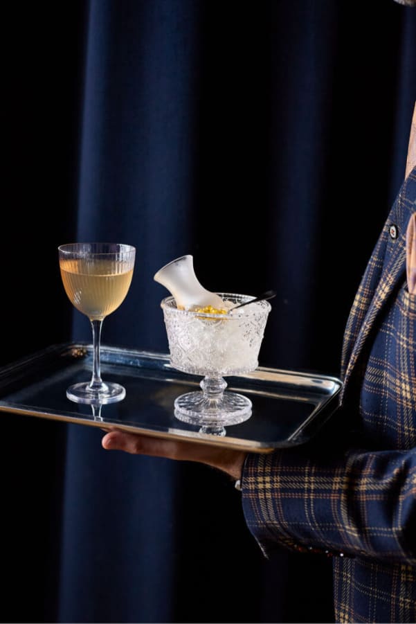 Server holding a tray with a cocktail glass and an ice-filled glass with a garnish in an elegant setting.